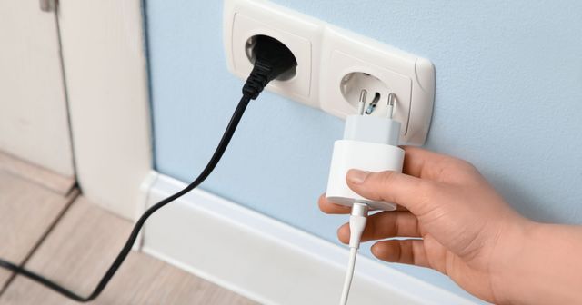 Een hand trekt een witte adapter uit een stopcontact in een lichtblauwe muur