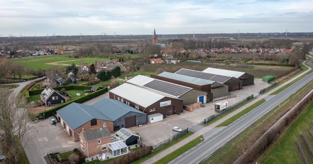 Luchtfoto van bedrijfsloodsen met zonnepanelen aan de rand van Nisse, met de dorpskerk op de achtergrond