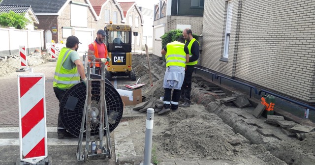 Wegwerkers in gele hesjes leggen kabels in een opengebroken trottoir naast een woonhuis